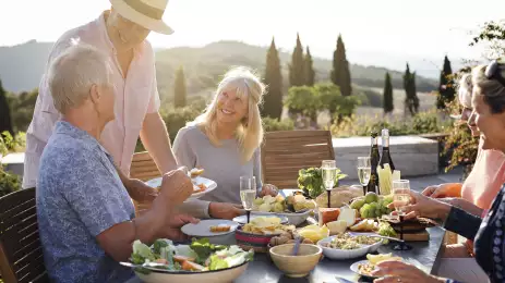 A group of mature friends are sitting around an outdoor dining table in Tuscany, Italy