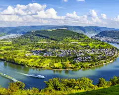 A wide shot of a bend in the Moselle River from it's surrounding hills under broad daylight, two river cruise ships can be seen creating gentle waves in the water.